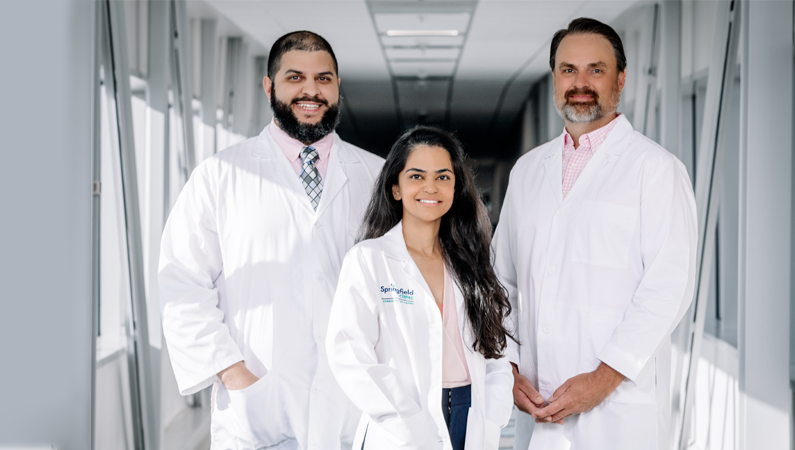 Three cancer doctors in white coats smiling in a naturally lit hallway.