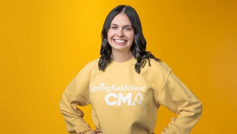 Young female with dark brown hair wearing yellow Springfield Clinic CMA top smiling in front of yellow background