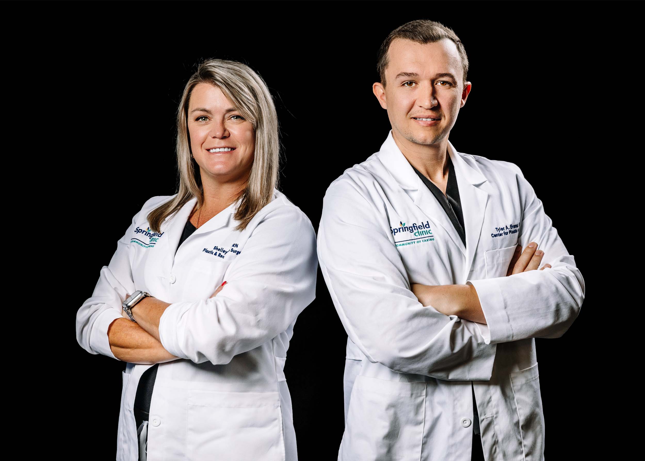 Male plastic surgery doctor smiling next to female nurse practitioner in white coats in front of black backdrop.