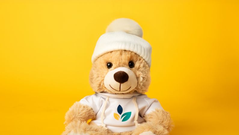 Stuffed animal bear wearing Springfield Clinic branded hoodie and white beanie posing in front of yellow background