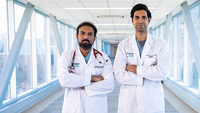 Two male doctors with dark hair standing with their hands crossed in a well-lit walkway