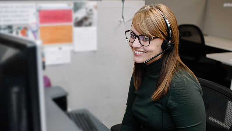 Female in office cubical wearing a headset smiling while looking at computer monitor.