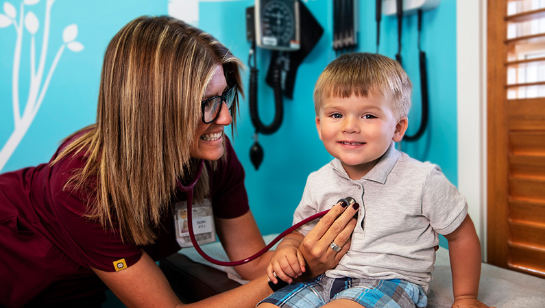 Child in bright blue exam room getting checked with stethoscope by smiling nurse