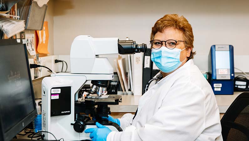 Laboratory technician in lab coat looking at camera with microscope.