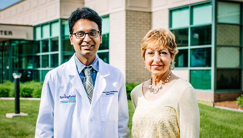 Male with dark hair wearing glasses and white doctor's coat standing next to female with short light hair in outdoor area