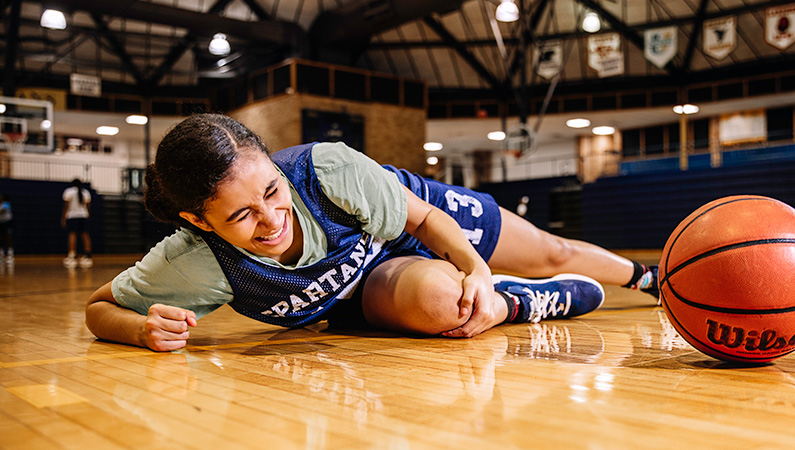 Young injured athlete laying on a indoor gym floor holding her knee in pain with a basketball in front of her.
