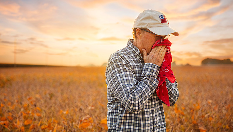 Male wearing hat sneezing into red handkerchief standing in front of freshly harvested field