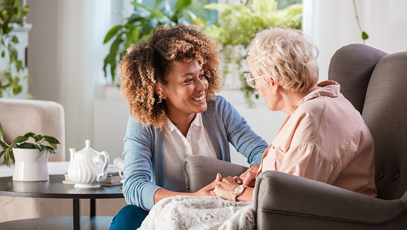Female with black curly hair sitting down holding hands with an elderly woman in an assisted living setting