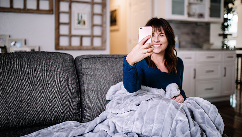 Young female in a home setting sitting on couch with fuzzy blanket looking into smartphone