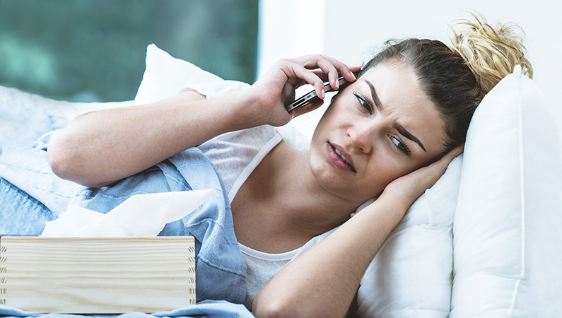 Young female laying in bed visibly uncomfortable with a box of tissues on the phone