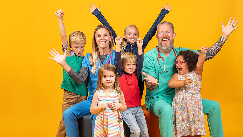 Two pediatric doctors celebrating with their hands up with 5 kids in front of a yellow backdrop.