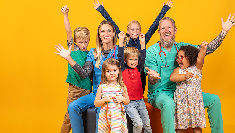 Two pediatric doctors celebrating with their hands up with 5 kids in front of a yellow backdrop.