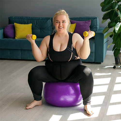 Female sitting on yoga exercise ball while holding 2 dumbbells in a living room setting.