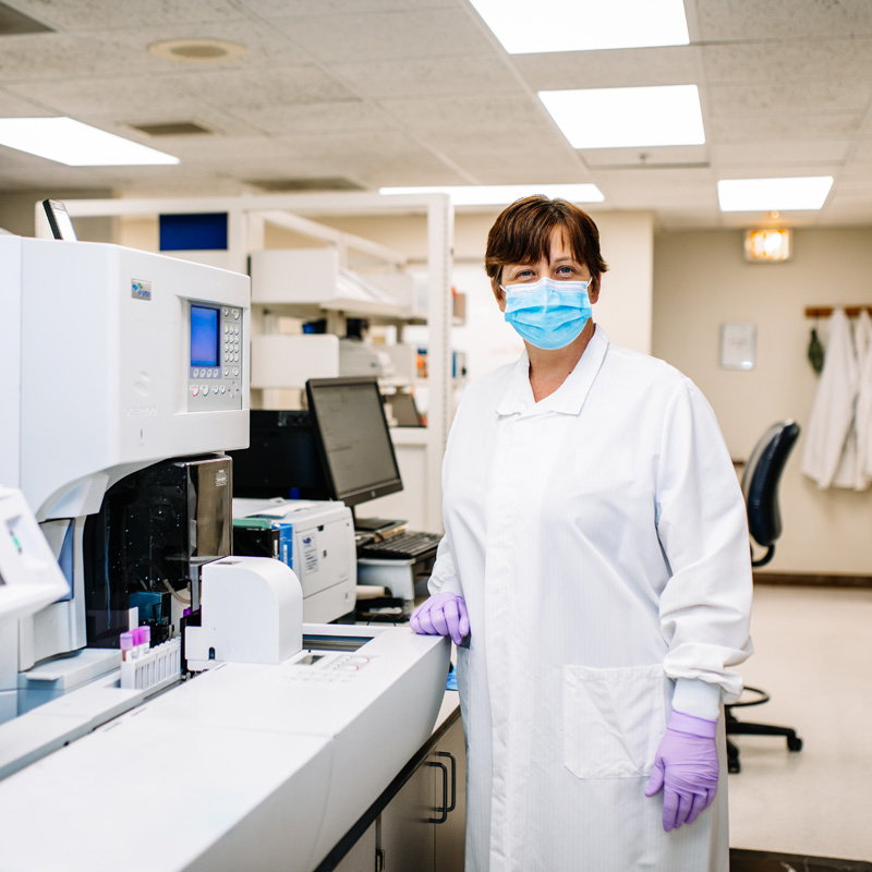 Lab technician in white lab coat, gloves and a mask looking at camera in laboratory.