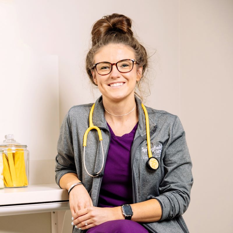 Female nurse sitting in patient exam room smiling at camera.