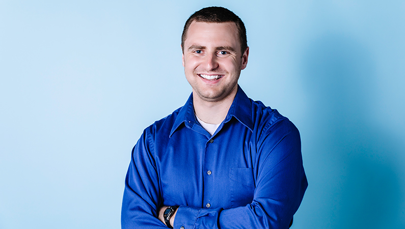 Male physical therapist with arms crossed smiling in front of a light blue backdrop.