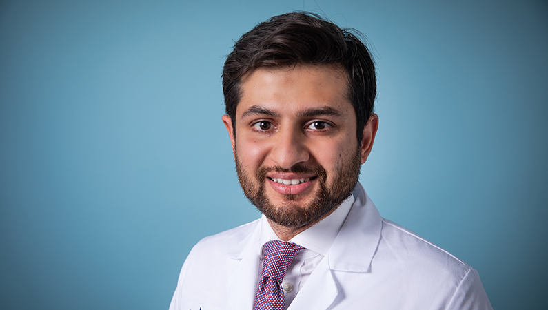 Male with dark hair wearing white medical coat smiling on studio-lit blue background