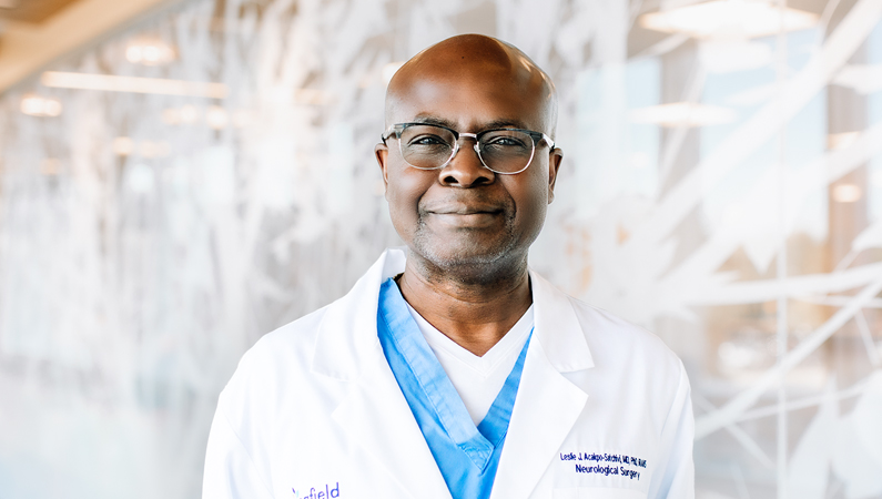 Male wearing glasses and blue scrubs with white medical coat posing in front of wall mural