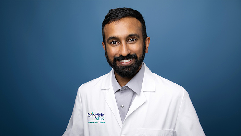 Male cardiologist wearing white medical coat while smiling in front of blue photo backdrop.