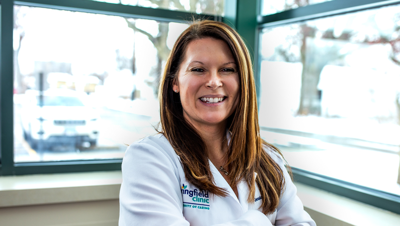 Female with dark straight hair wearing white medical coat smiling in front of windows