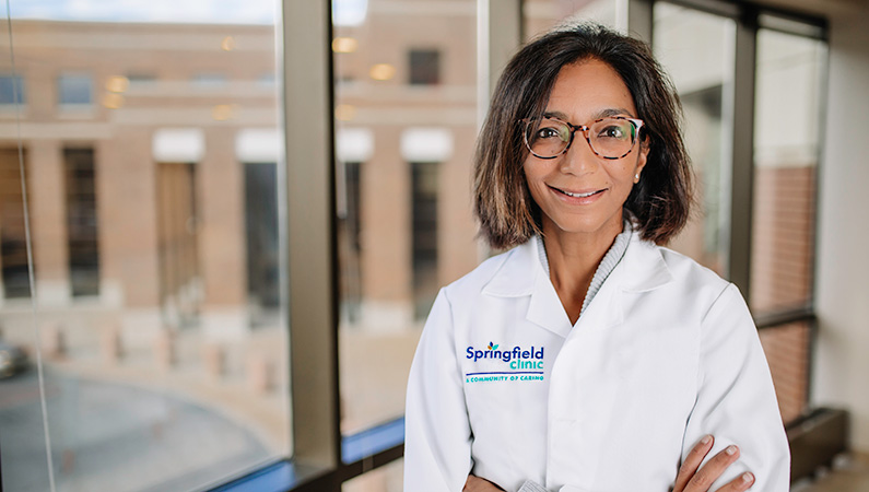 Woman wearing a white doctors coat smiling in front of window.