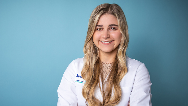 Young female with long blonde hair smiling in white coat against bright blue background
