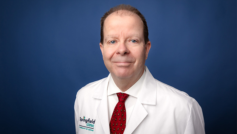 Male physician assistant in white medical coat smiling in front of blue photo backdrop.
