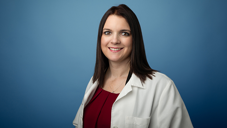 Female nurse practitioner wearing white medical coat smiling in front of blue backdrop.