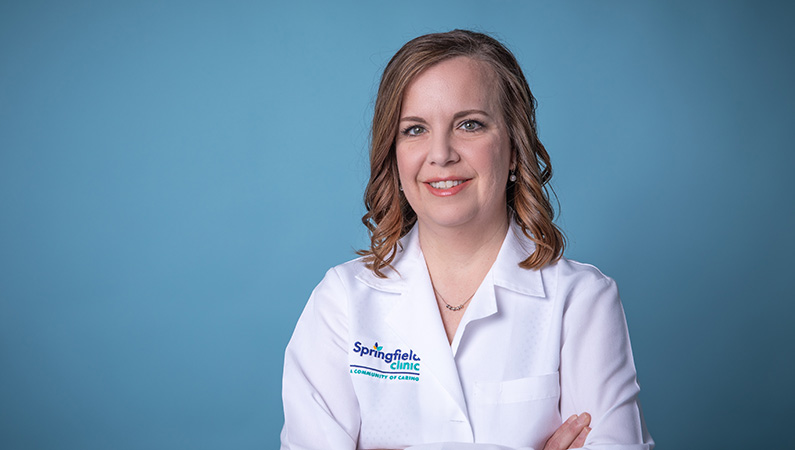 Female with short hair wearing white medical coat smiling in front of studio-lit blue background