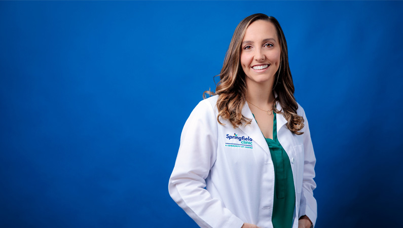 Female internal medicine doctor in white lab coat smiling in front of a royal blue backdrop.