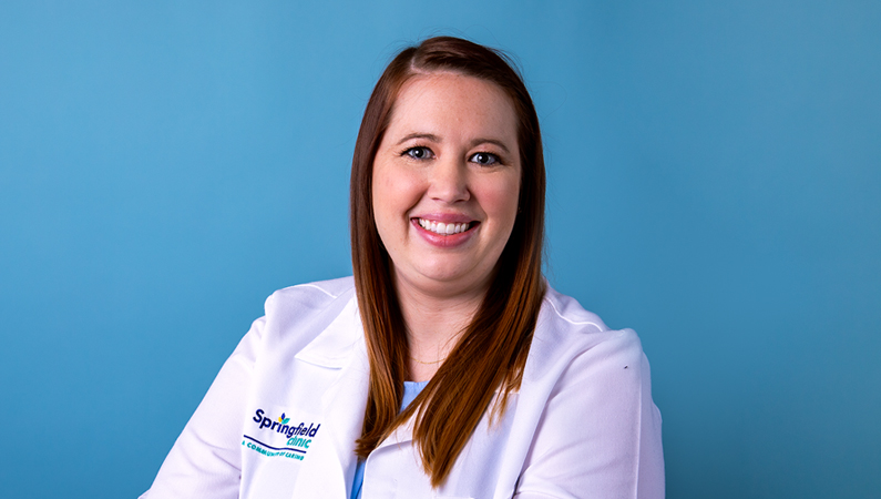 Female with dark hair wearing white medical coat smiling in front of bright blue background