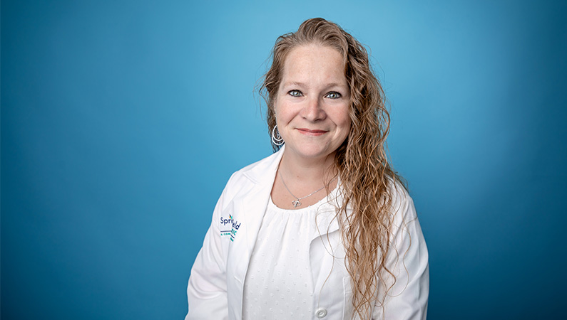 Female nurse practitioner in white coat smiling in front of light blue backdrop.