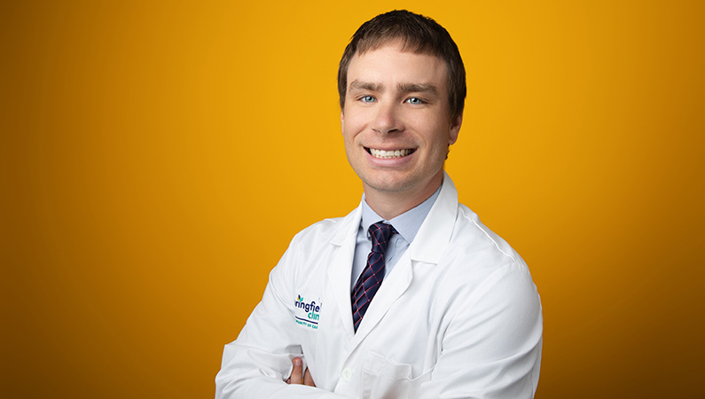 Male physician wearing white medical coat smiling in front of yellow photo backdrop.