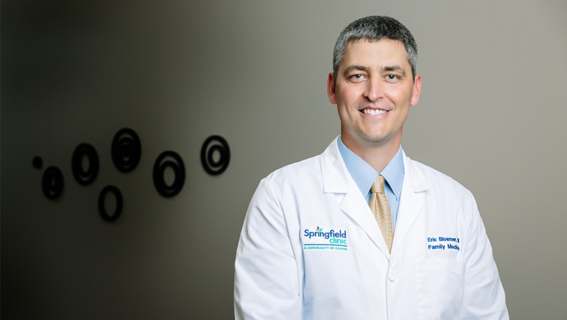 Male family medicine doctor in white coat smiling in a dim patient hallway.