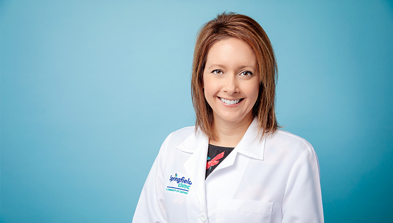 Female nurse practitioner smiling in white coat in front of light blue photo backdrop.