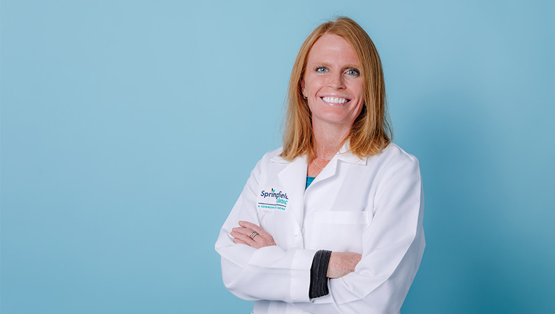 Female nurse practitioner posing in front of light blue backdrop in white coat.