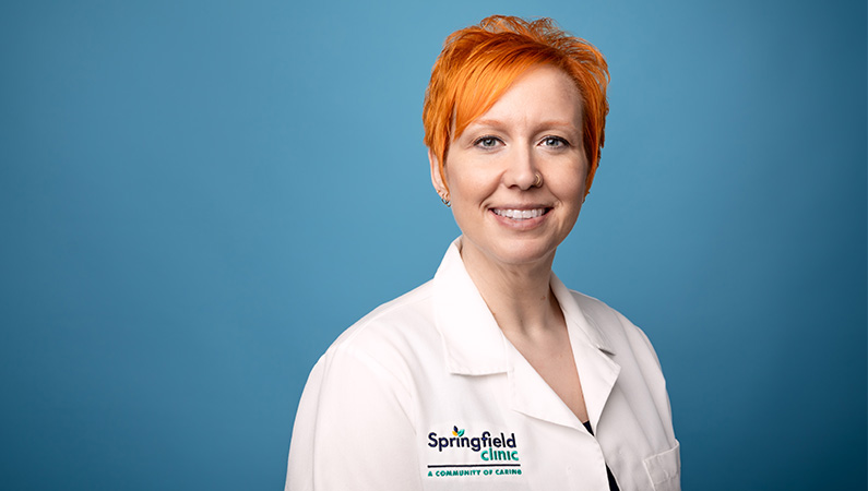 Female nurse wearing white medical coat smiling in front of a light blue backdrop.