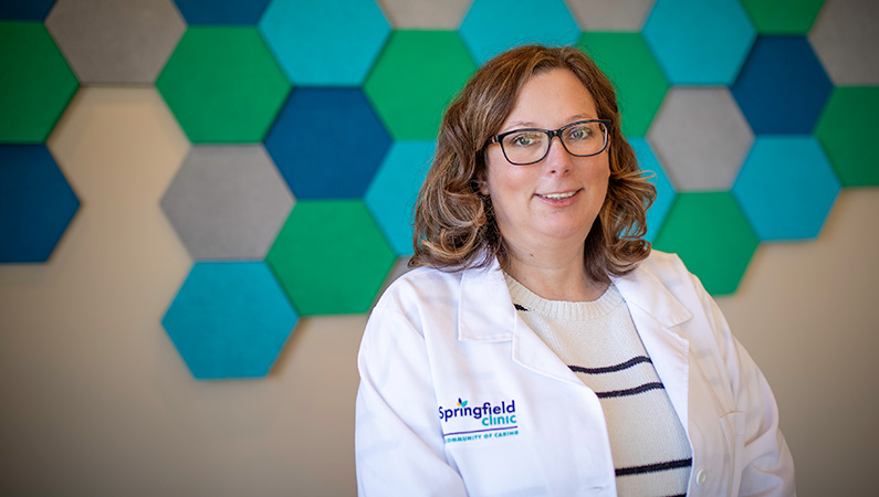 Woman in doctors coat smiling in front of a wall with green and blue hexagonal decorations.