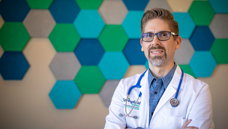 Man in doctors coat smiling in front of a wall with green and blue hexagonal decorations.