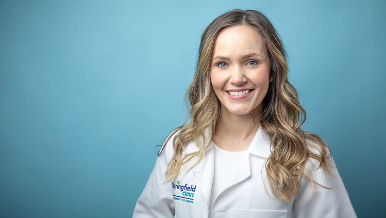 Female physician assistant in white lab coat smiling in front of a light blue photo backdrop.