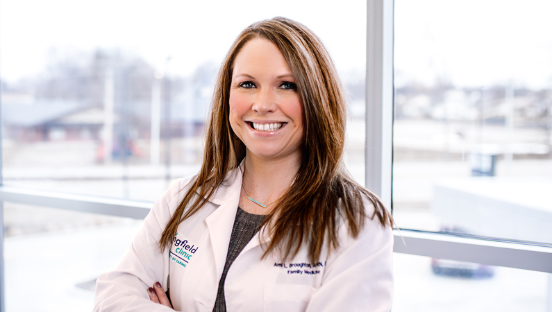 Female with dark straight hair wearing white medical coat smiling in front of windows