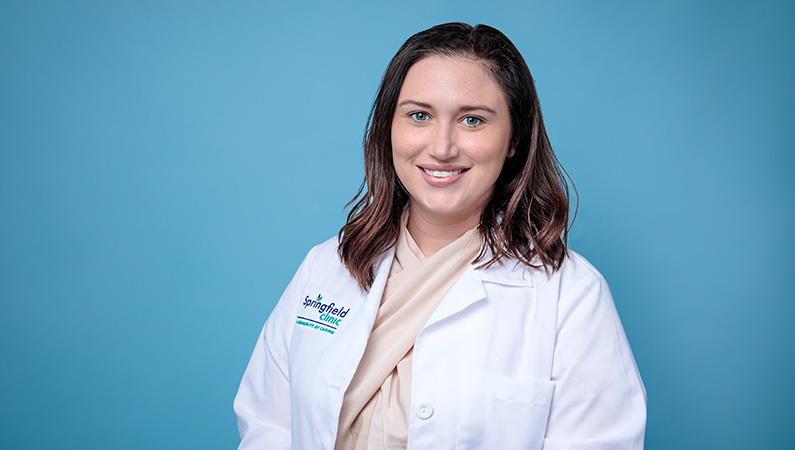 Female nurse practitioner in white coat smiling in front of light blue backdrop.