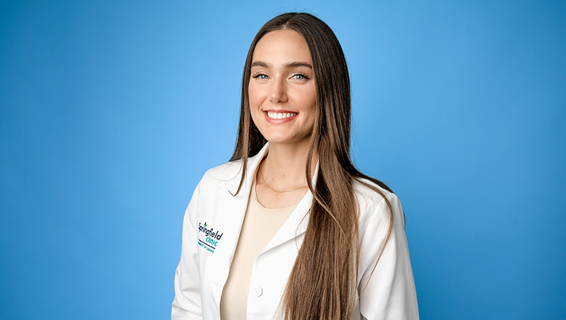 Female physician assistant in white lab coat smiling in front of a light blue photo backdrop.