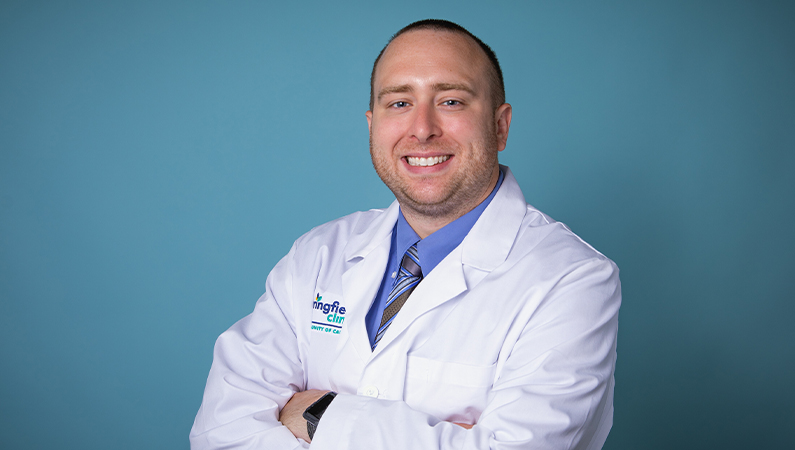 Male nurse practitioner in white coat smiling in front of a teal blue backdrop with his arms crossed.