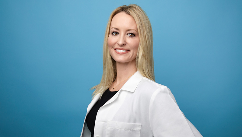 Female nurse practitioner in white medical coat smiling in front of blue photo backdrop.