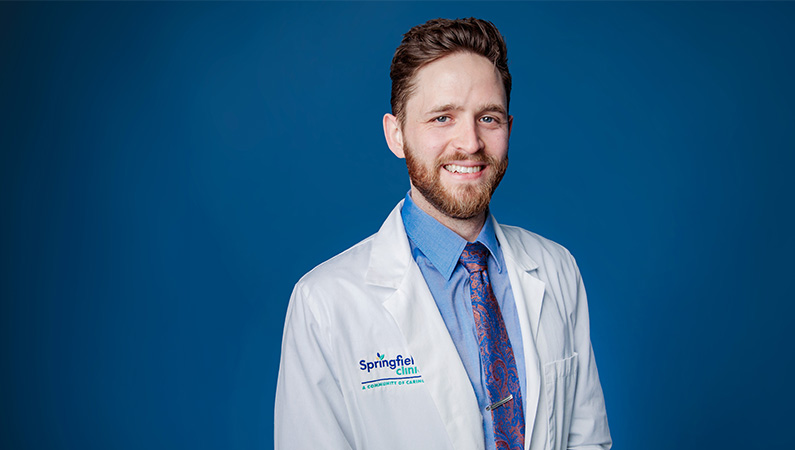 Male neurologist smiling in front of light blue backdrop.