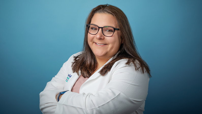 Female nurse practitioner with brown hair wearing white medical coat smiling in front of blue photo backdrop.