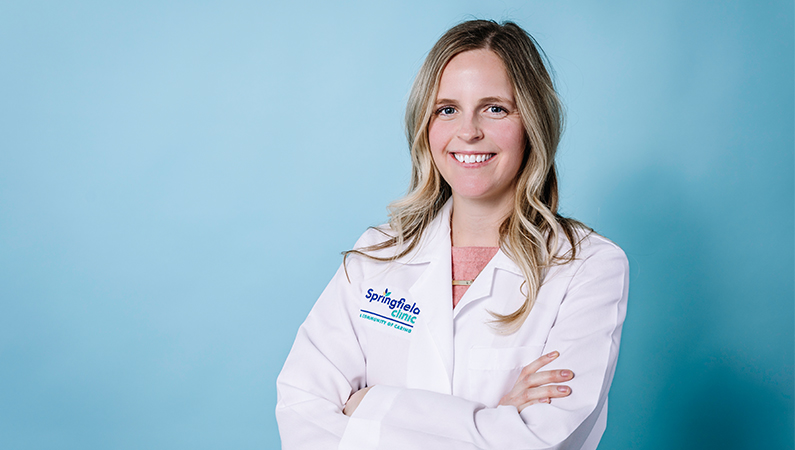 Female nurse practitioner wearing white coat smiling in front of a light blue backdrop.