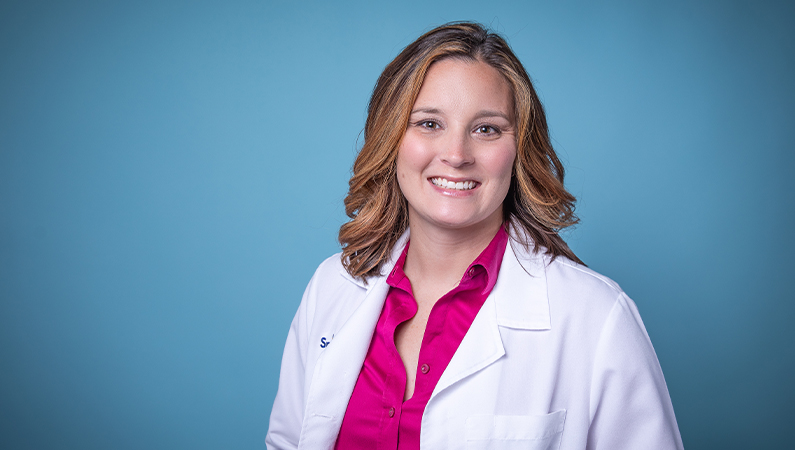 Female nurse practitioner in white coat smiling in front of a light blue backdrop.