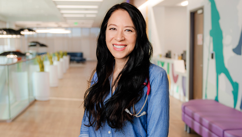 Female pediatrician with long dark hair smiling in a clinical setting.